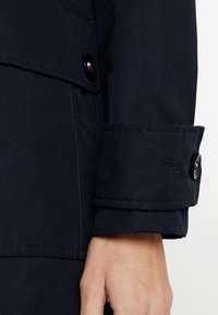 Trench coat blu navy con una texture morbida, chiusura con bottoni e logo, e risvolto con iniziali ricamate.