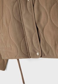 Gros plan sur une veste marron matelassée montrant un surpiqûre en forme de vague, une fermeture à boutons-pression et un cordon de serrage réglable à l'ourlet.