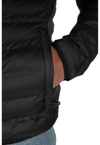 Schwarze Puffy-Jacke mit gestepptem Muster, seitlichem Taschen und elastischen Bündchen. Das Material wirkt glänzend und glatt, geeignet für kaltes Wetter.