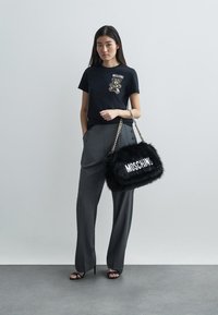 Juoda marškinėliai su lokio grafika ir "Moschino" logotipu, derinami su pilkais plačiakomečiais kelnais, laikant juodą dirbtinio kailio rankinę su balta užrašu.