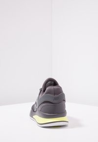 Šedá sportovní obuv s elegantním designem, textilním svrškem, polstrovaným límcem a žlutým akcentem na podrážce. Na patě je logo a boty mají texturované povrchy.