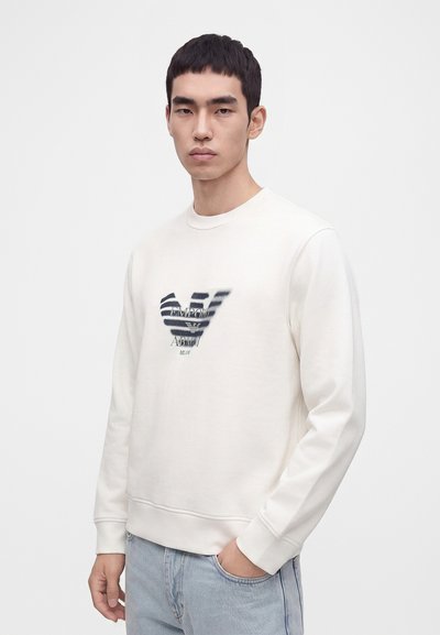Jeune homme aux cheveux courts et foncés portant un sweat-shirt blanc avec un logo noir flou et un jean bleu clair, debout devant un fond uni.