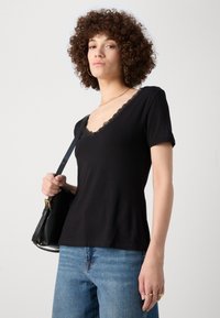 Svart t-skjorte med korte ermer og V-hals i blondekant, kombinert med blå vide jeans og en svart håndveske. Teksturen virker myk.