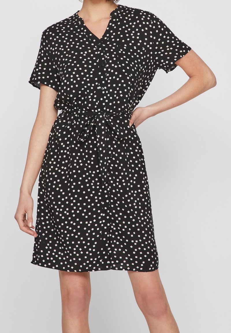 Femme portant une robe noire à manches courtes avec des motifs carrés blancs, devant boutonné et ceinture nouée à la taille.