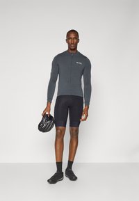 Maillot de cyclisme à manches longues gris foncé avec une fermeture éclair à l'avant, cuissard rembourré noir, chaussures de cyclisme noires et un casque noir tenu d'une main.