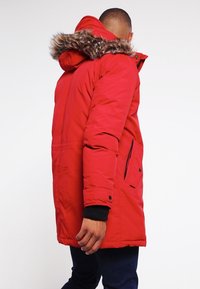 Parka isolée rouge avec une capuche doublée de fourrure, dotée d'une fermeture éclair à l'avant, de poches latérales et de poignets noirs. Tissu texturé avec une finition mate.