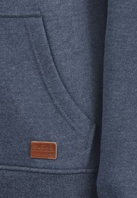 Sudadera azul marino con un tejido texturizado, que presenta un bolsillo frontal y una etiqueta de cuero marcada con "BLEND" cosida cerca del dobladillo.