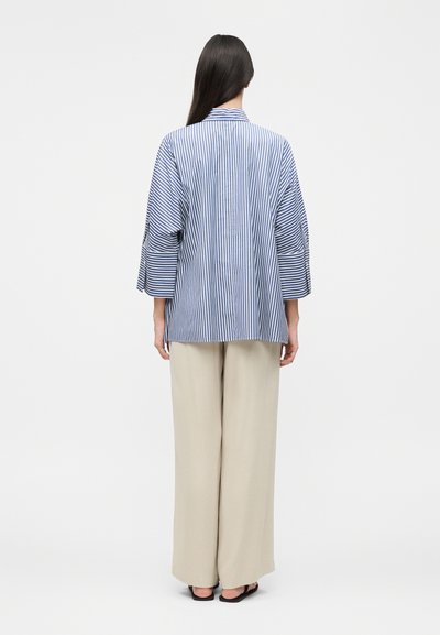 Γυναίκα με μακριά σκούρα μαλλιά στέκεται με την πλάτη, φοράει μπλε και λευκή κάθετα ριγέ μπλούζα με φαρδιά μπεζ παντελόνια και μαύρα σανδάλια.