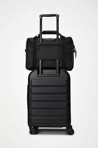 Sort hard-shell koffert med horisontale riller og fire hjul, med en svart stoffduffelbag på toppen som har en frontlomme med glidelås og stropper.