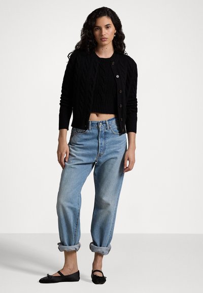 Cardigan negru tricotat cu model, purtat peste un pulover negru scurt; blugi de culoare albastru deschis, cu talie înaltă și manșete răsucite; pantofi joși negri cu detaliu sub formă de fundiță.