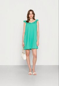 Vestido verde sem mangas com decote franzido e um corte solto. Combinado com uma bolsa branca e sandálias planas. Textura de tecido suave.