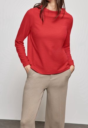 Frau trägt ein rotes Langarmshirt und beige Hose, steht mit den Händen in den Taschen vor einem schlichten hellen Hintergrund.