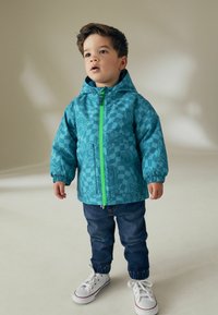 Blå mönstrad barnjacka med huva, med en grön frontdragkedja och elastiska manschetter. Bärs över mörka jeansbyxor och vita sneakers.
