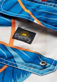 Camisa colorida azul y blanca con patrones de hojas, que presenta un acento naranja, una etiqueta que dice "Superdry" y un detalle de botón de metal a presión.