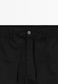 Black pants waistband with elastic band, belt loops, button closure, and tied drawstring front detail.