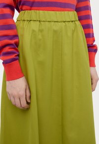 Gonna verde a pieghe con elastico in vita, abbinata a un maglione a maniche lunghe a strisce rosse e viola, mostrata sul torso e sulla mano di un modello.