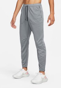 Vīrietis, kurš valkā gaiši pelēkas sašaurinātas sporta bikses ar priekšējām kabatām un baltas sporta kurpes ar rozā akcentiem, stāvot pret baltu fona.