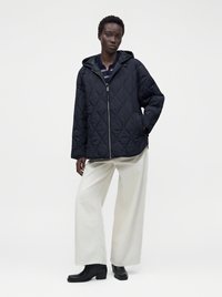 Personne portant une veste matelassée noire à capuche, un pantalon blanc à jambes larges, des bottines noires, debout devant un fond clair uni.