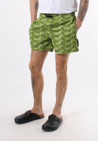 Costume da bagno verde con motivo di testo nero ripetuto, vita elastica e cordoncino regolabile. Accompagnato da zoccoli neri senza lacci.