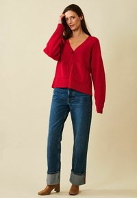 Kvinna som bär en röd cardigan med V-ringning och framfickor, uppvikta blå jeans och beige ankelboots med klack, stående mot en enkel beige bakgrund.