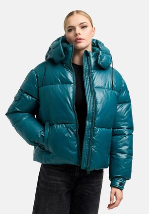 Frau mit blondem Haar, die eine petrolfarbene Steppjacke mit Kapuze, ein schwarzes Oberteil und dunkle Jeans vor einem unifarbenen Hintergrund trägt.