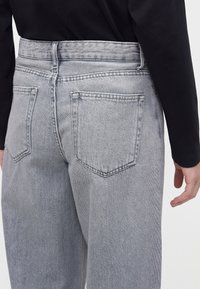 Personne portant un jean en denim gris clair taille haute avec deux poches arrière et un haut noir à manches longues, main détendue sur le côté.