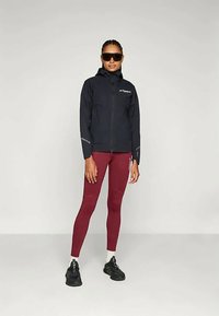 Črna vodoodporna jakna s kapuco in odsevnimi detajli, v kombinaciji z burgundskimi legicami in črnimi športnimi čevlji. Model nosi sončna očala.