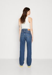 Vaqueros de denim azul de cintura alta con piernas anchas, caracterizados por un lavado claro y dos bolsillos traseros. La modelo lleva una camiseta sin mangas blanca.