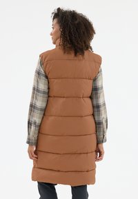 Bruine puffer vest met een gewatteerd ontwerp, schouderflappen en zijsplitten, gecombineerd met een geruite, lange mouwen shirt eronder.