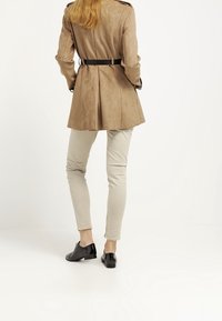 Brązowy zamszowy płaszcz trencz, dopasowana sylwetka, z naramiennikami. W połączeniu z jasnobeżowymi dopasowanymi spodniami i czarnymi płaskimi butami.