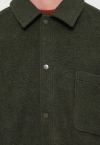 Oliivivihreä villapaita-takki, jossa on kaulus, etunapit ja timanttitasku. Ominaisuuksiin kuuluu pehmeä tekstuuri ja huomaamattomat tikkauslinjat.