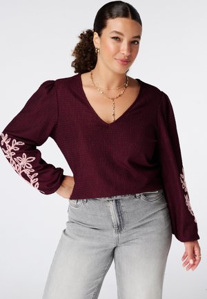 Femme aux cheveux bouclés portant un pull bordeaux avec une broderie florale rose sur les manches et un jean gris, posant avec la main sur la hanche.