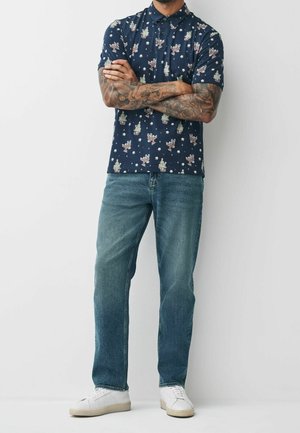 Mand stående med armene krydset iført en mørkeblå kortærmet skjorte med tegneserieudskrifter, blå jeans, hvide sneakers og armtatoveringer.