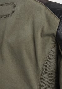 Dos d'une veste verte avec un patch d'épaule foncé, couture détaillée autour du col et zone matelassée texturée près de la couture latérale.