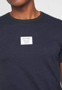 Tummansininen puuvillapaita, jossa on erottuva tumma kaulus, varustettuna suorakulmaisella kangastarralla, jossa lukee "Jack & Jones" ja tuotekuvaukset.