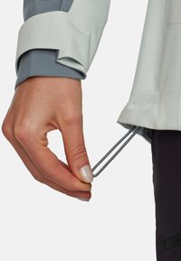 Håndjusterer snørebåndet i kanten af en grå jakke, der bæres over mørke bukser, med jakkens ærme delvist dækende håndleddet.