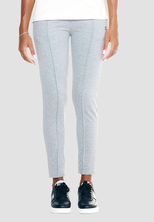 Persoon draagt slim-fit lichtgrijze broek en zwarte sneakers, staand tegen een effen lichte achtergrond.