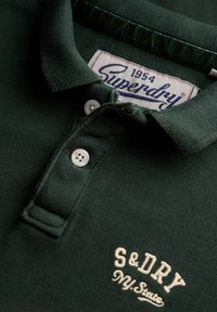 Donkergroen poloshirt met een gestructureerde stof, twee witte knopen en een geborduurd logo in gebroken wit. Label met de tekst "Superdry 1954".