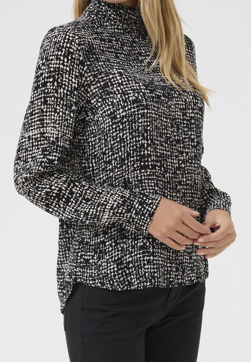 Femme portant un chemisier à manches longues à motifs noir et blanc avec un col montant, associé à un pantalon noir, les mains doucement jointes.