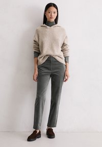Beżowy, puszysty sweatshirt z dekoltem w serek i długimi rękawami, w zestawieniu z ciemnoszarymi spodniami z krótkiego weluru i brązowymi butami wsuwanymi. Gładka struktura na całej powierzchni.