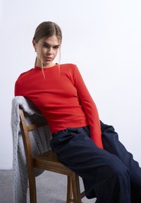 Νεαρή γυναίκα με μακριά μαλλιά δεμένα πίσω, φοράει κόκκινο μακρυμάνικο μπλουζάκι και σκούρο φαρδύ παντελόνι, σκύβει πίσω σε μια ξύλινη καρέκλα με μια γκρι ζακέτα απλωμένη πάνω της.