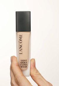 Botella de base de vidrio esmerilado con liquididad beige en su interior y una tapa cuadrada negra elegante. El texto en la botella incluye "Lancôme" y "Teint Idole Ultra Wear."
