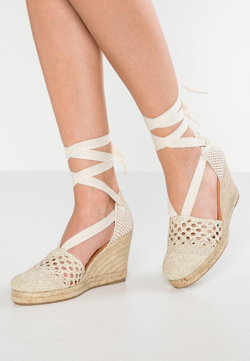 Jambes portant des espadrilles compensées en beige tissé avec des brides qui s'attachent à la cheville, debout sur un sol lisse de couleur claire.
