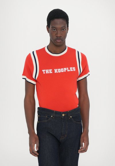 Punainen lyhythihainen t-paita, jossa on valkoisia ja laivastonsinisiä raitoja, ja jossa on "THE KOOPLES" -logo valkoisena rinnan kohdalla. Yhdistetty tummansinisiin farkkuihin.