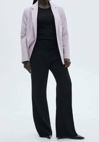 Veste légèrement lavande portée sur un haut noir côtelé et un pantalon large noir, présentant des lignes épurées et des chaussures noires pointues.