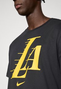 Musta puuvillainen t-paita, jossa suuri keltainen "LA"-kuvio edessä. Pyöreä kaula-aukko ja lyhyet hihat ovat näkyvissä.