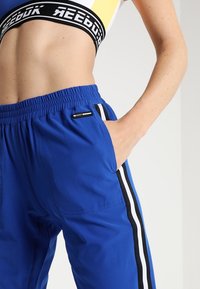 Blå sportbyxor med svarta och vita sidostreck, elastisk midja och fickor. Materialet verkar slätt och lätt.