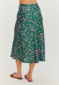 Zielona satynowa spódnica z różowym wzorem przypominającym liście, sylwetka w linii A, detal w pasie oraz długość do połowy łydki. Noszone płaskie sandały.