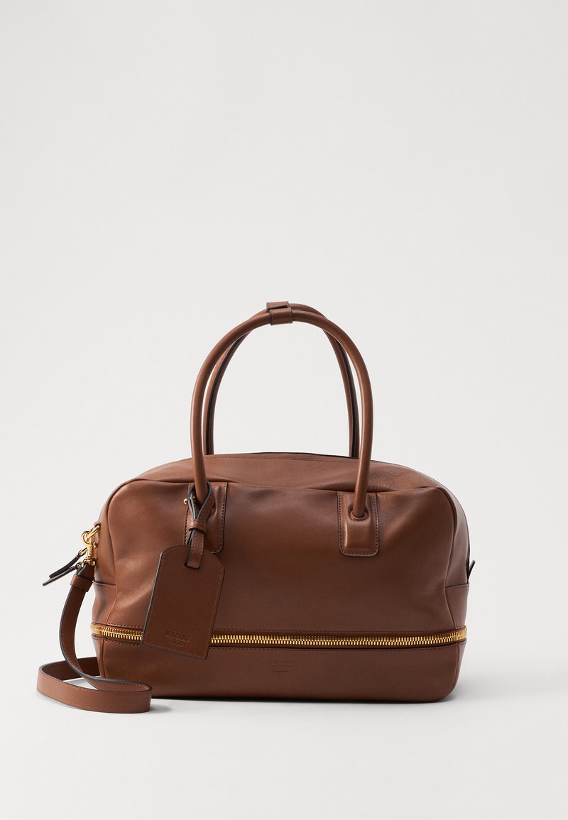 Sac de voyage en cuir marron avec fermeture éclair dorée, poignées supérieures doubles, bandoulière amovible et une étiquette de bagage attachée. Texture lisse.