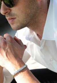 Bracciale in metallo argentato con design a catena, superfici lucidate e accenti circolari. Indossato su un polso, sopra una camicia di colore chiaro.